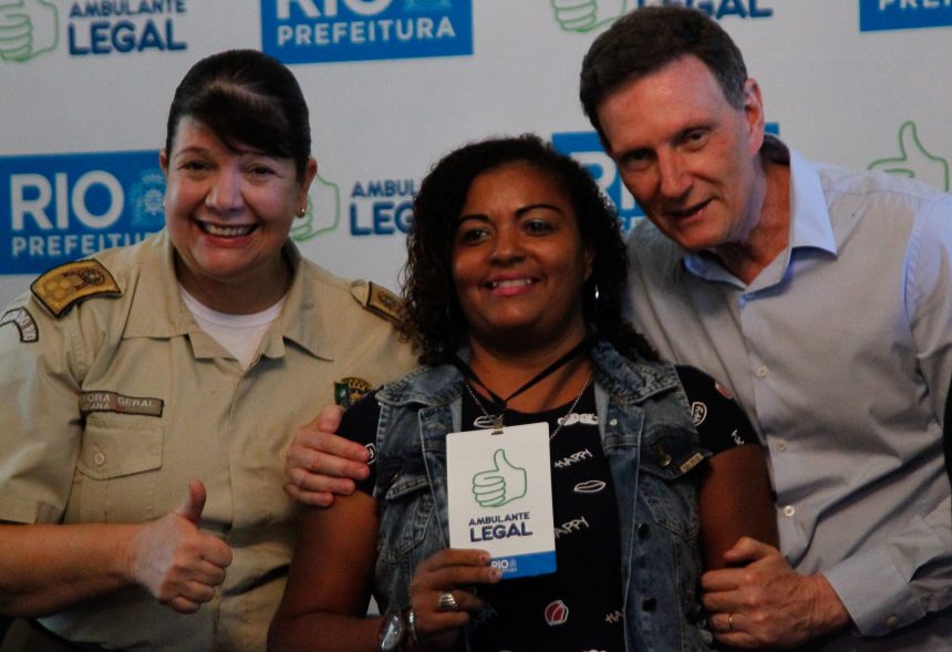 O prefeito Crivella entrega crachá a ambulante legalizada da Zona Oeste, ao lado da inspetora Tatiana Mendes, comandantes da Guarda Municipal