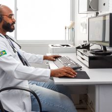 Médico de óculos e barba, sentado com jaleco branco em frente ao computador ,fazendo o atendimento através da teleconsulta